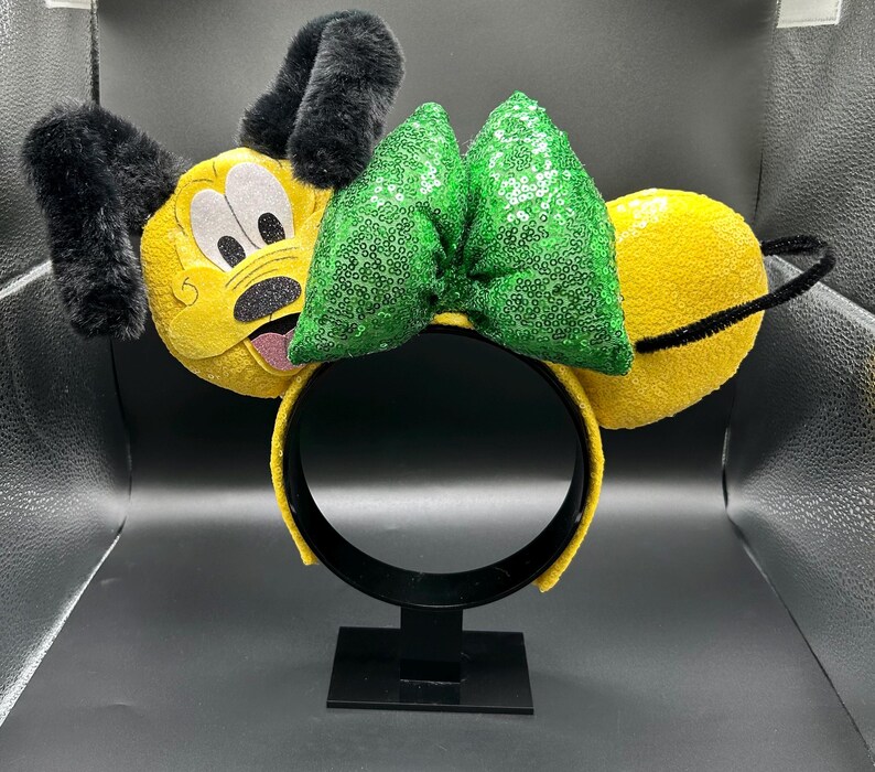 May include: A yellow headband with plush dog ears and a green sequined bow. The dog's face is embroidered on the front of the headband, with black ears and a black nose. The headband is on a black stand.