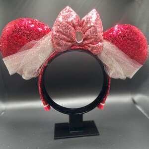 Op de afbeelding: Rode en roze Minnie Mouse oren met pailletten, met een witte tule strik en een heldere strasssteen in het midden van de strik. De oren zitten op een zwarte hoofdband.