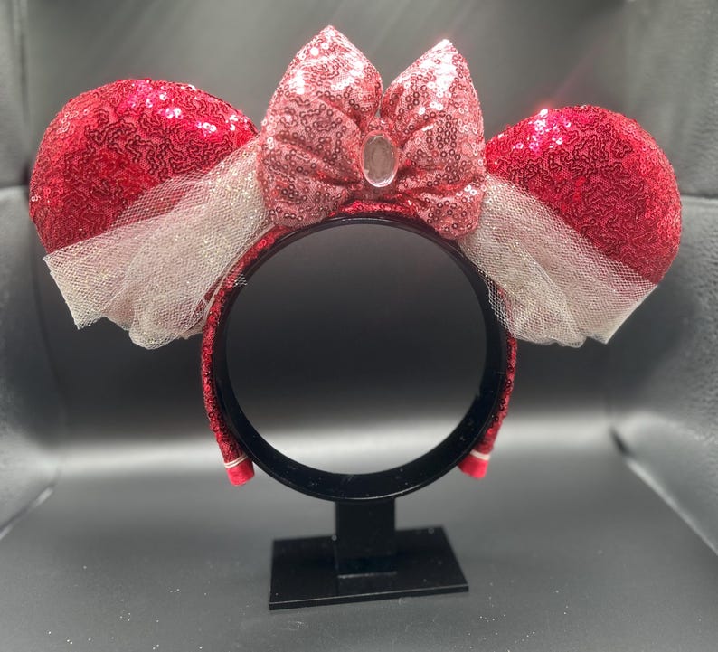 Op de afbeelding: Rode en roze Minnie Mouse oren met pailletten, met een witte tule strik en een heldere strasssteen in het midden van de strik. De oren zitten op een zwarte hoofdband.