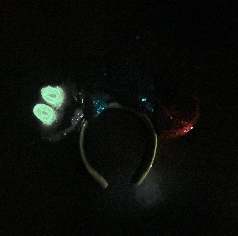May include: A black headband with two glowing green circular shapes on the left side and a red sequined bow on the right. The headband has blue sequins and a dark background.