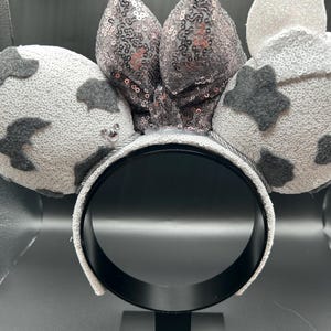 May include: A headband featuring two round ears covered in white sequins with dark grey felt cow-print patches. A central bow made of shimmering silver sequins sits between the ears. The headband frame is wrapped in matching white sequins.