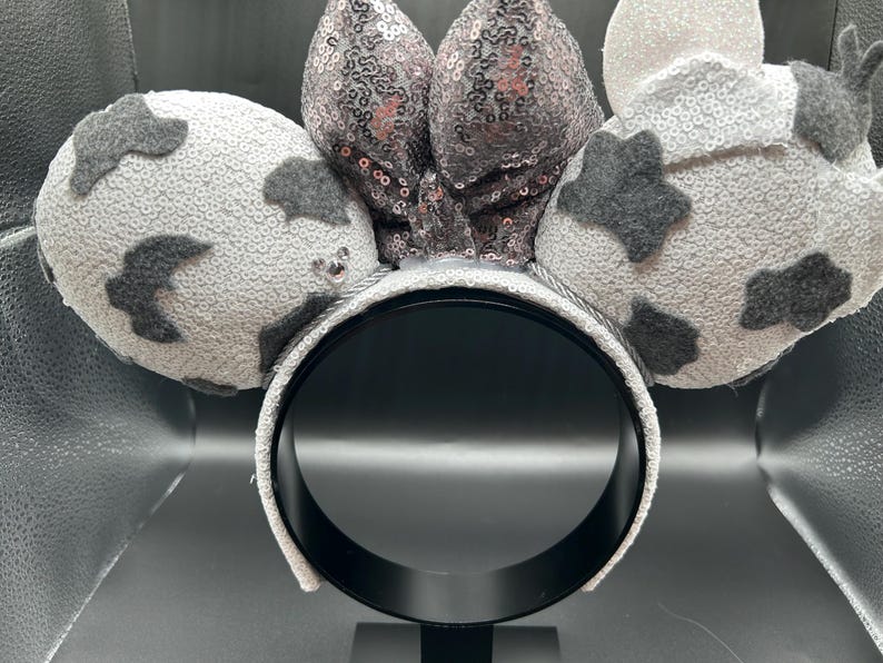 May include: A headband featuring two round ears covered in white sequins with dark grey felt cow-print patches. A central bow made of shimmering silver sequins sits between the ears. The headband frame is wrapped in matching white sequins.