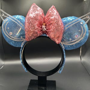 May include: A decorative headband with blue sequined ears and a pink sequined bow. The headband has a black band and is adorned with silver accents and clear embellishments. A small bottle is attached to the bow.