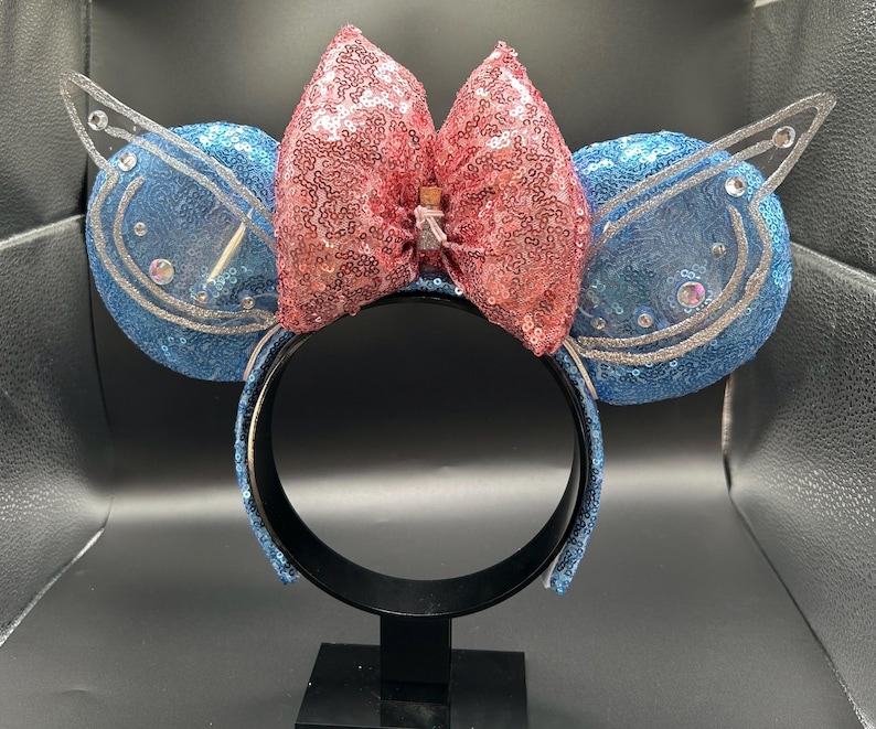 May include: A whimsical headband featuring blue sequined ears with silver accents and a pink sequined bow. The headband has a black band and is decorated with small, clear embellishments. A small bottle is attached to the bow.