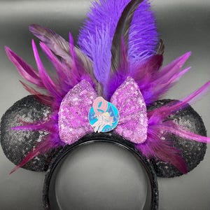 May include: A headband featuring two black sequined ears, a central light purple sequined bow, and a cluster of vibrant purple and dark violet feathers. A circular enamel pin depicting a stylized character is centered on the bow.