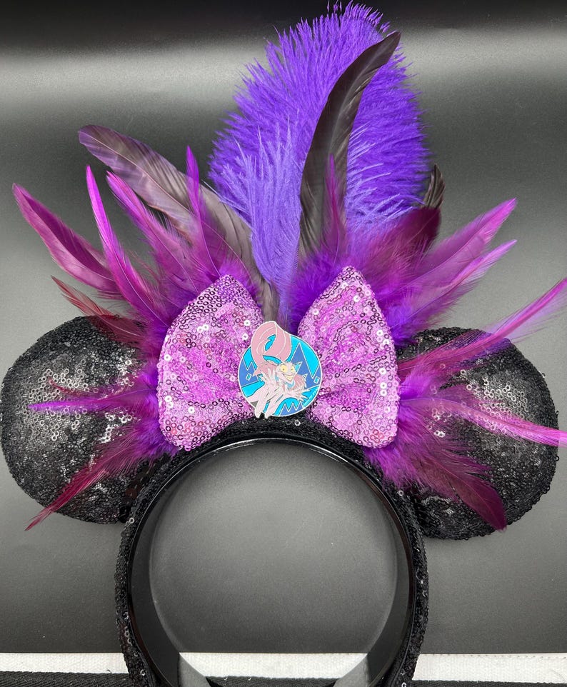 May include: A headband featuring two black sequined ears, a central light purple sequined bow, and a cluster of vibrant purple and dark violet feathers. A circular enamel pin depicting a stylized character is centered on the bow.
