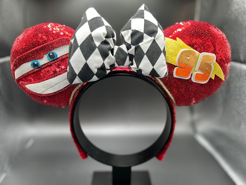 May include: Red and black Mickey Mouse ears with a checkered bow, a lightning bolt, and the number 95 on one ear and a cartoon face on the other ear.