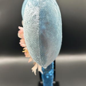 Peut inclure: Une vue lat&eacute;rale d'un serre-t&ecirc;te avec de grandes oreilles arrondies recouvertes de velours bleu clair. Le serre-t&ecirc;te est orn&eacute; d'un bouquet de fleurs artificielles rose p&acirc;le et blanches sur le c&ocirc;t&eacute;. L'arceau est recouvert de sequins bleus.