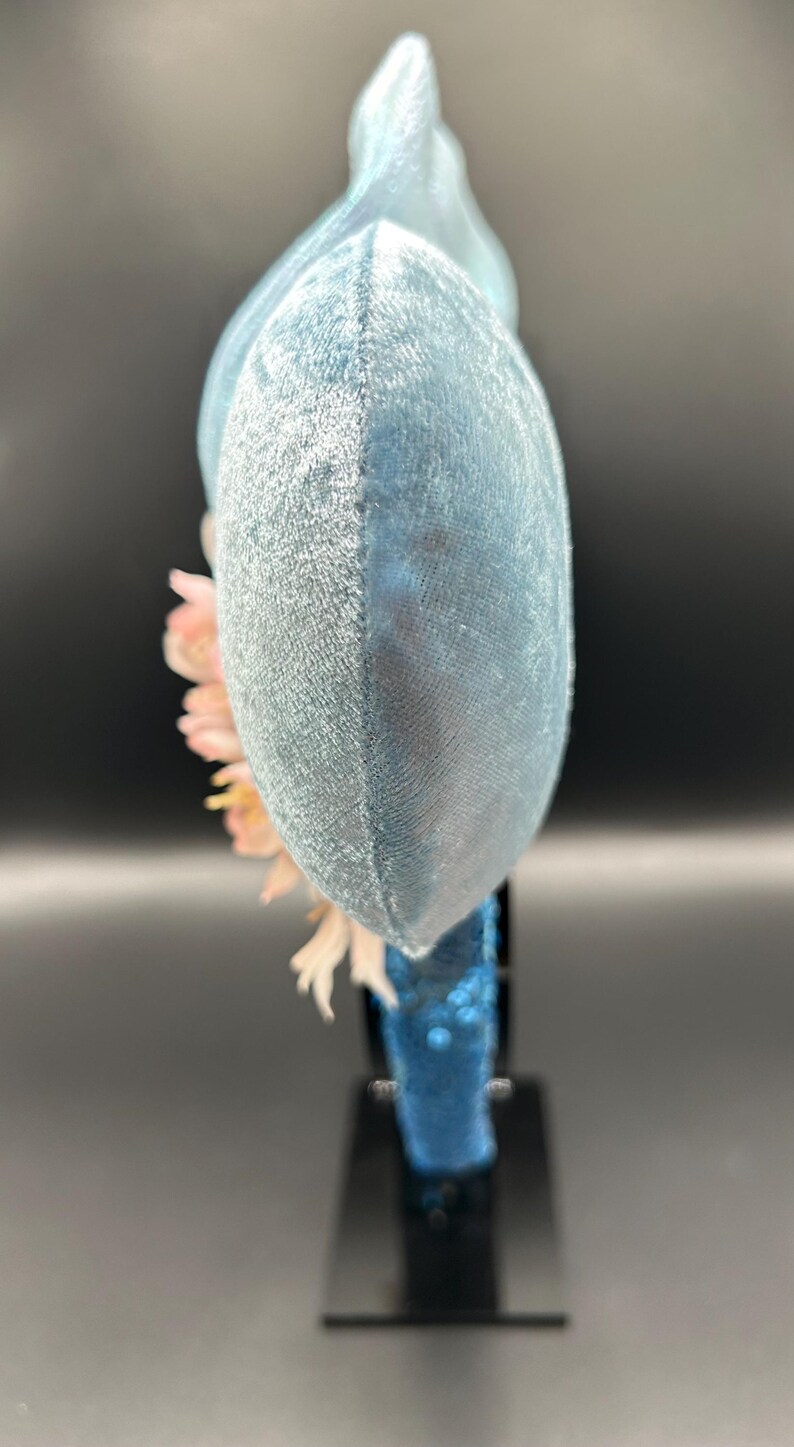 Peut inclure: Une vue lat&eacute;rale d'un serre-t&ecirc;te avec de grandes oreilles arrondies recouvertes de velours bleu clair. Le serre-t&ecirc;te est orn&eacute; d'un bouquet de fleurs artificielles rose p&acirc;le et blanches sur le c&ocirc;t&eacute;. L'arceau est recouvert de sequins bleus.