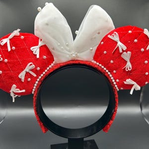 May include: Red and white Minnie Mouse ear headband. The red ears are quilted and adorned with white bows and pearl accents. The white bow is layered with pearls. The headband is black with red sequins and a pearl trim.