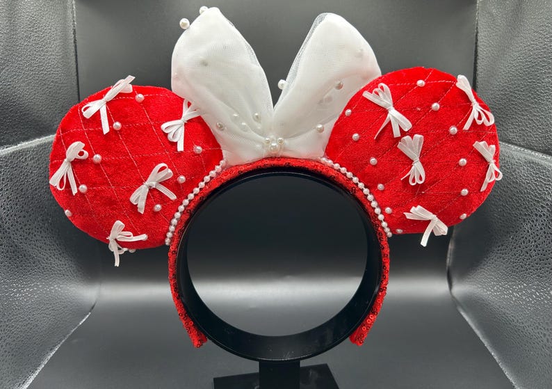 May include: Red and white Minnie Mouse ear headband. The red ears are quilted and adorned with white bows and pearl accents. The white bow is layered with pearls. The headband is black with red sequins and a pearl trim.