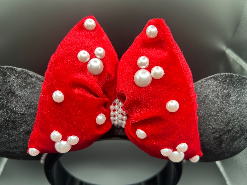 K&ouml;nnte beinhalten: Rotes Samt-Minnie-Maus-Ohrenband mit einer gro&szlig;en Schleife, die mit wei&szlig;en Perlen verziert ist. Die Schleife hat zwei Mickey-Maus-Formen aus Perlen.