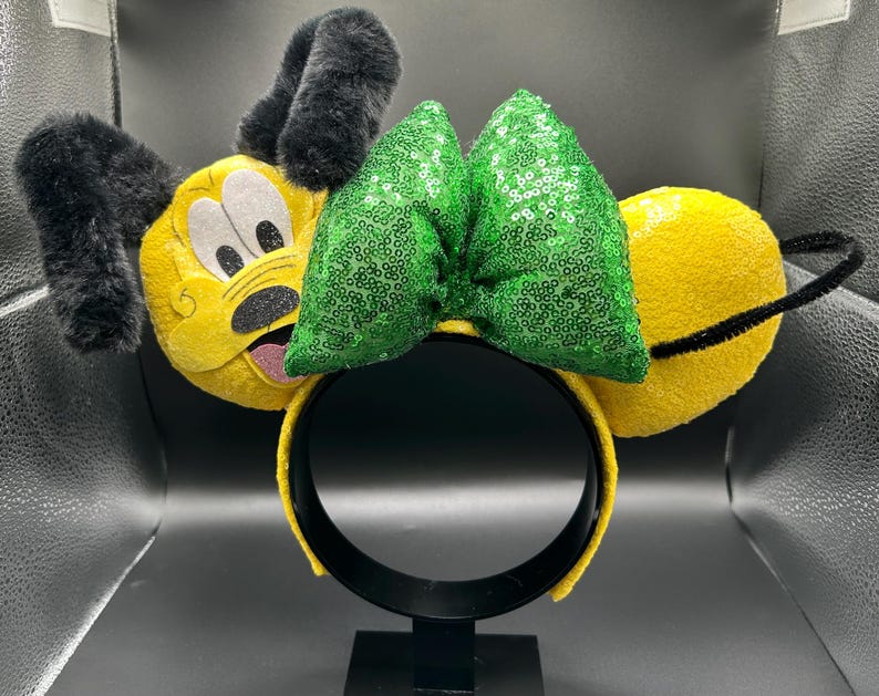 May include: Yellow character ears with black fuzzy ears and a green sequined bow. The headband is yellow and black. The character's face is yellow with black and white details.