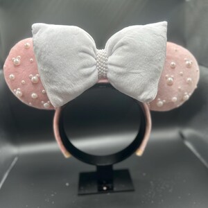 May include: Pink mouse ears headband with a white bow and pearl embellishments.