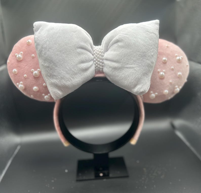 May include: Pink mouse ears headband with a white bow and pearl embellishments.