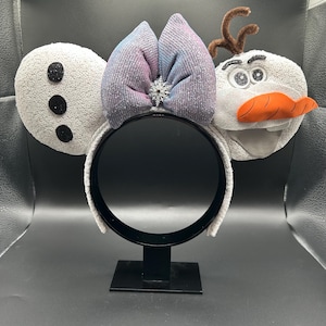 May include: A whimsical headband featuring Olaf from Frozen. The ears are white with black accents, a bow with pink and blue hues, and a snowflake center. Olaf's face has an orange carrot nose and twig hair.