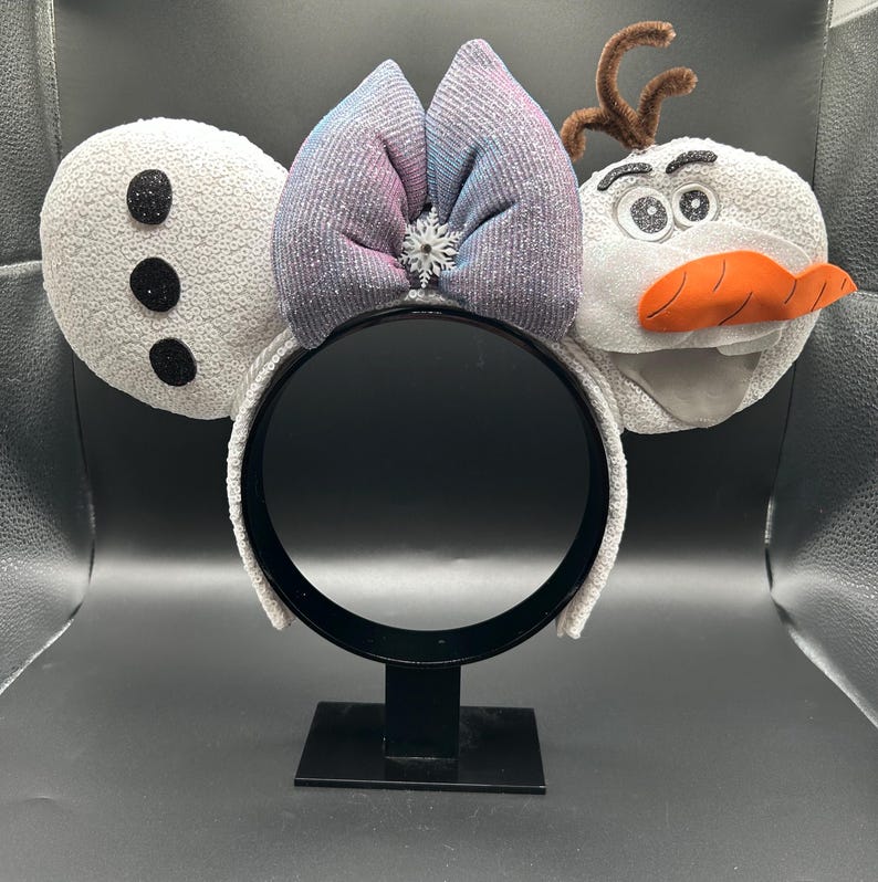 May include: A whimsical headband featuring Olaf from Frozen. The ears are white with black accents, a bow with pink and blue hues, and a snowflake center. Olaf's face has an orange carrot nose and twig hair.