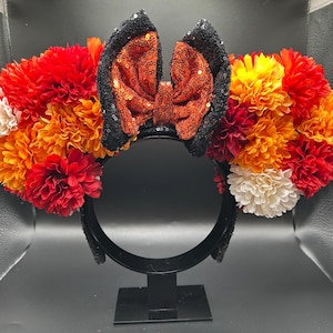 May include: A headband featuring two ears adorned with a dense arrangement of artificial flowers in shades of red, orange, yellow, and cream. Between the ears sits a central bow covered in shimmering orange sequins with black sequined edges.