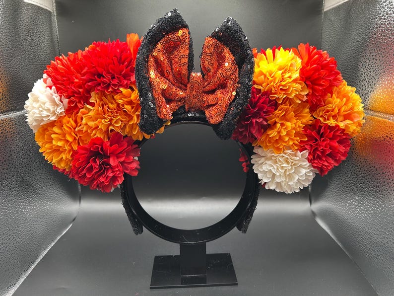 May include: A headband featuring two ears adorned with a dense arrangement of artificial flowers in shades of red, orange, yellow, and cream. Between the ears sits a central bow covered in shimmering orange sequins with black sequined edges.