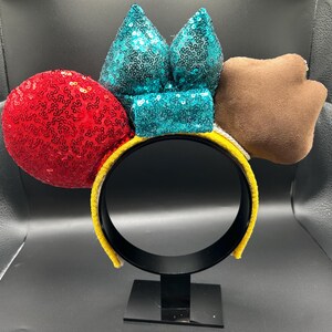May include: A yellow headband with a black interior, featuring a red sequined sphere, teal sequined bow, and a brown paw-shaped accent. The headband is designed for wear and is displayed on a black stand.