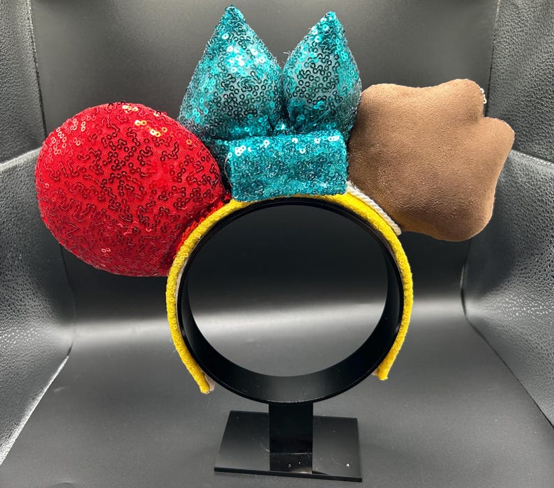May include: A yellow headband with a black interior, featuring a red sequined sphere, teal sequined bow, and a brown paw-shaped accent. The headband is designed for wear and is displayed on a black stand.
