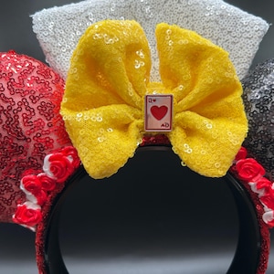 May include: A headband featuring two circular ears covered in red and black sequins. A large yellow lace bow sits in the center, adorned with a small rectangular charm depicting a Queen of Hearts playing card. White sequins form a background element.