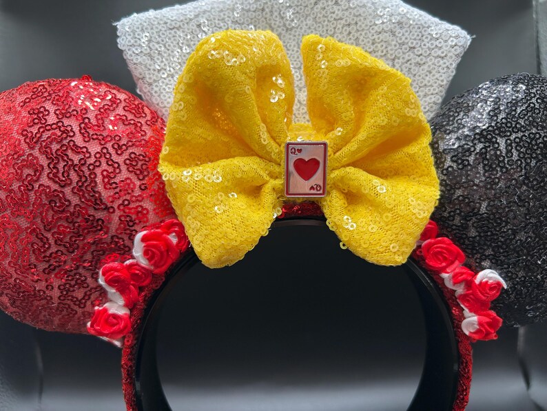 May include: A headband featuring two circular ears covered in red and black sequins. A large yellow lace bow sits in the center, adorned with a small rectangular charm depicting a Queen of Hearts playing card. White sequins form a background element.