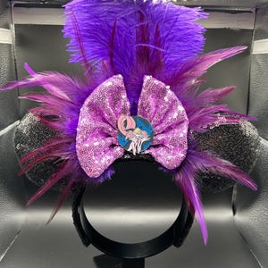 May include: A headband featuring two black sequined ears, a large pink sequined bow, and vibrant purple feathers. A circular pin depicting a pink creature with large eyes and sharp teeth is centered on the bow, surrounded by dark purple plumage.