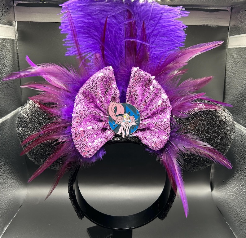 May include: A headband featuring two black sequined ears, a large pink sequined bow, and vibrant purple feathers. A circular pin depicting a pink creature with large eyes and sharp teeth is centered on the bow, surrounded by dark purple plumage.