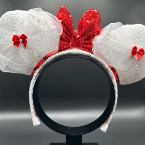 May include: White Minnie Mouse ears with red bows and a red sequin bow. The ears are made of white tulle and sequins.