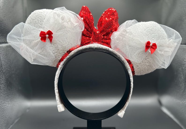 May include: White Minnie Mouse ears with red bows and a red sequin bow. The ears are made of white tulle and sequins.