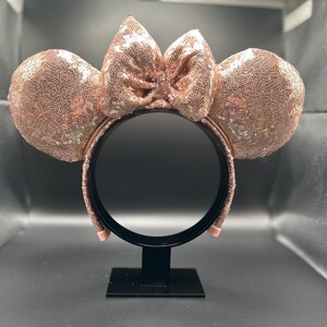 May include: Rose gold sequined mouse ear headband with a matching bow. The ears and bow are covered in shimmering sequins. The headband is black and has a rose gold band. A fun accessory for themed events.