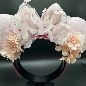 May include: A light pink mouse ear headband with plush ears. The headband features a large bow made of white tulle, decorated with white flowers and pink rhinestones. Clusters of pink and white flowers are also around the ears.