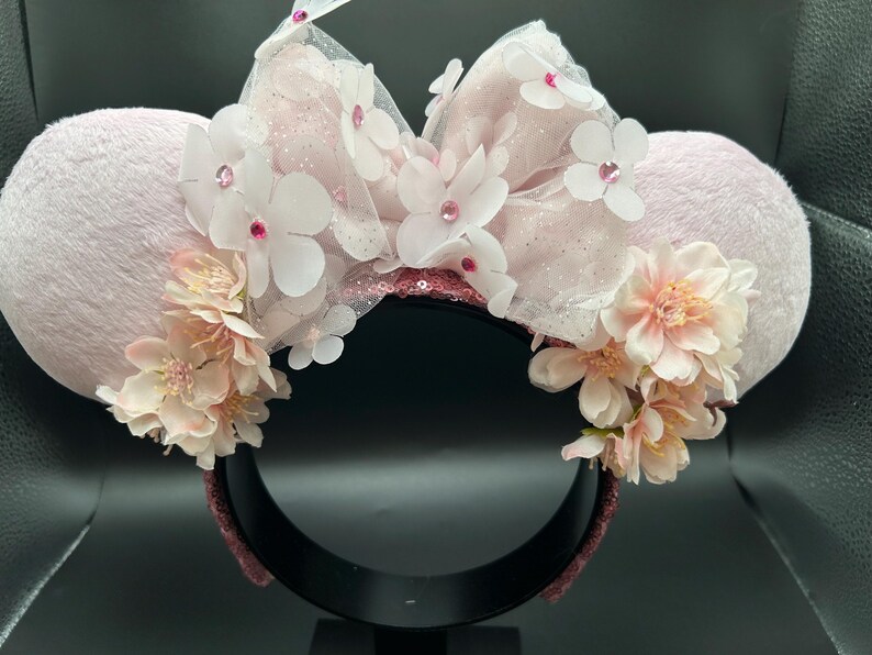 May include: A light pink mouse ear headband with plush ears. The headband features a large bow made of white tulle, decorated with white flowers and pink rhinestones. Clusters of pink and white flowers are also around the ears.