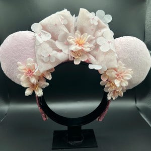 Op de afbeelding: Een haarband met twee pluizige lichtroze ronde oren en een grote centrale strik. De strik is versierd met gelaagde witte en lichtroze bloemapplicaties en kleine kraaltjes. Extra trossen lichtroze bloemen zijn bevestigd aan de basis van elk oor, wat een bloemstuk cre&euml;ert aan de voorzijde van de haarband.