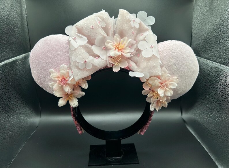 Op de afbeelding: Een haarband met twee pluizige lichtroze ronde oren en een grote centrale strik. De strik is versierd met gelaagde witte en lichtroze bloemapplicaties en kleine kraaltjes. Extra trossen lichtroze bloemen zijn bevestigd aan de basis van elk oor, wat een bloemstuk cre&euml;ert aan de voorzijde van de haarband.