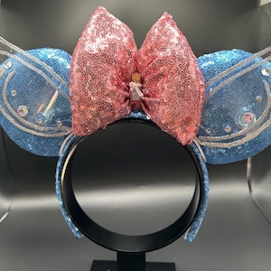 May include: A blue sequined headband with two round blue ears, each with a clear plastic wing. A large pink sequined bow sits between the ears. The headband is black.