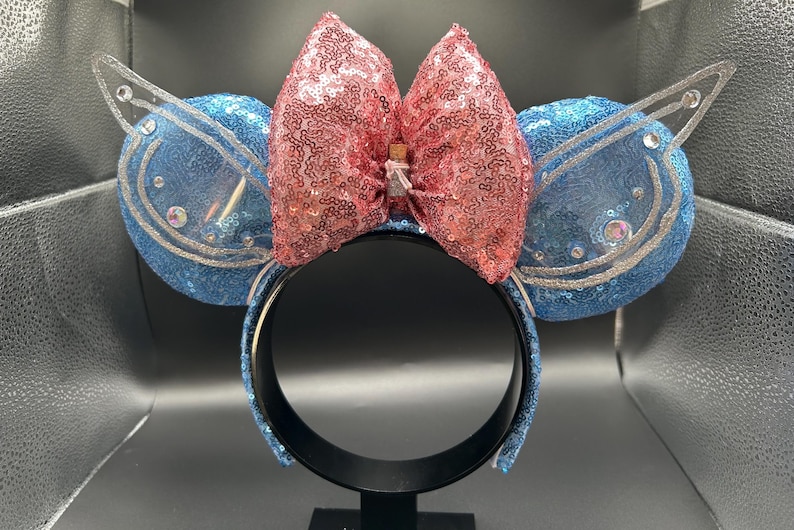 May include: A blue sequined headband with two round blue ears, each with a clear plastic wing. A large pink sequined bow sits between the ears. The headband is black.