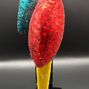 May include: A decorative headband with two sequined ornaments. One ornament is red, the other is teal. The headband base is yellow. The items are displayed on a black stand against a black background.