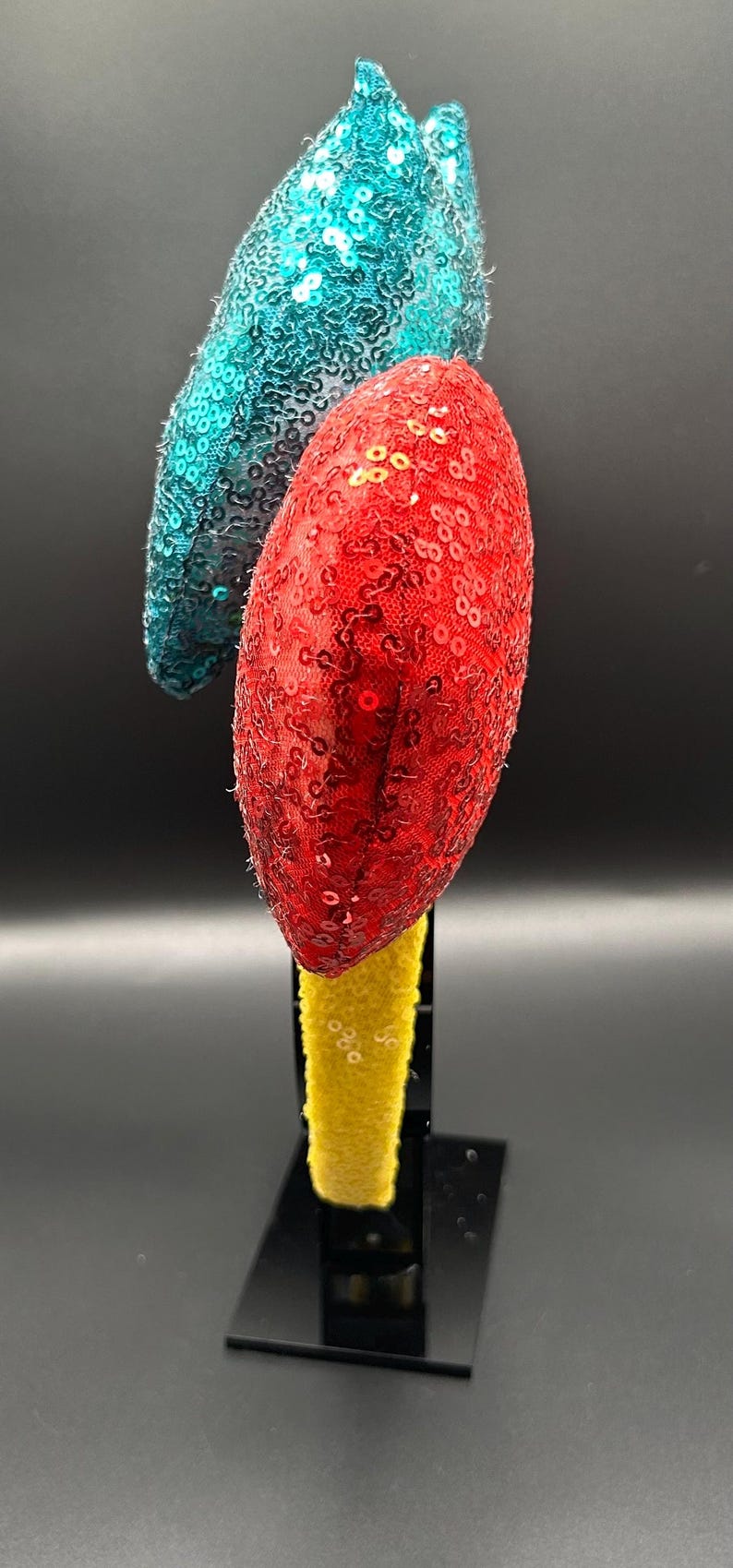 May include: A decorative headband with two sequined ornaments. One ornament is red, the other is teal. The headband base is yellow. The items are displayed on a black stand against a black background.