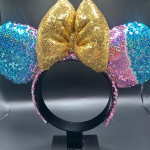 May include: A headband featuring two round ears covered in blue, pink, and purple sequins. A large, central bow made of gold sequins sits between the ears. The headband frame is black, and the inner band is lined with pink sequins.