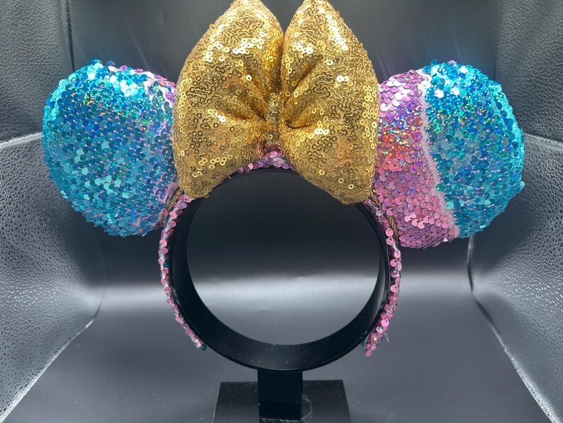 May include: A headband featuring two round ears covered in blue, pink, and purple sequins. A large, central bow made of gold sequins sits between the ears. The headband frame is black, and the inner band is lined with pink sequins.