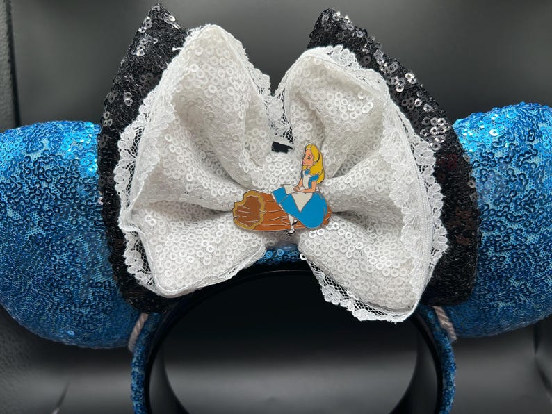 May include: Blue sequined mouse ears with a white sequined bow and black sequined accents. A small pin of Alice from Alice in Wonderland is attached to the bow. The headband is black.