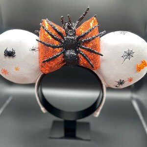 May include: Halloween-themed headband with white plush ears decorated with spiderwebs and spiders. A sequined orange bow with a black glitter spider is centered on the headband. A festive accessory for Halloween.