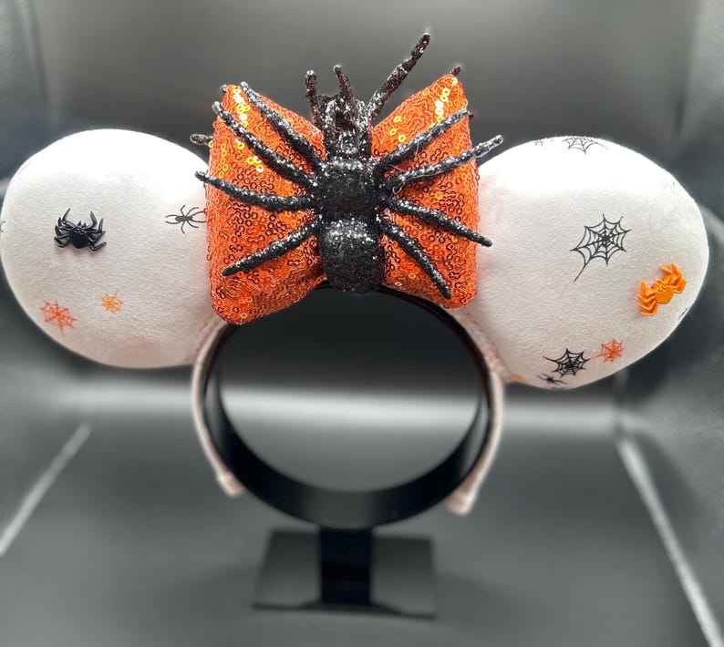 May include: Halloween-themed headband with white plush ears decorated with spiderwebs and spiders. A sequined orange bow with a black glitter spider is centered on the headband. A festive accessory for Halloween.
