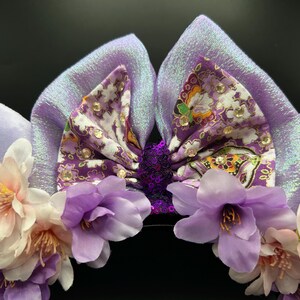 Puede incluir: Un par de orejas de color lavanda con un lazo central de estampado floral morado y pedrer&iacute;a transparente. El lazo tiene un centro de lentejuelas moradas oscuras. La base est&aacute; adornada con flores artificiales de color lila y blanco.