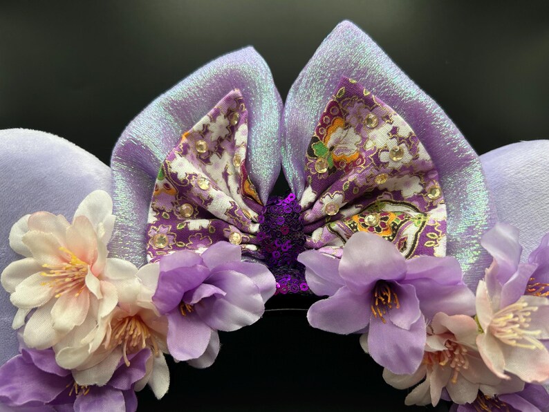 Puede incluir: Un par de orejas de color lavanda con un lazo central de estampado floral morado y pedrer&iacute;a transparente. El lazo tiene un centro de lentejuelas moradas oscuras. La base est&aacute; adornada con flores artificiales de color lila y blanco.