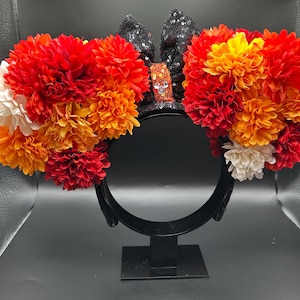 May include: A headband featuring two large circular clusters of artificial flowers in shades of red, orange, and white. Between the floral clusters sits a black sequined bow with a central orange sequined knot featuring a small silver icon.