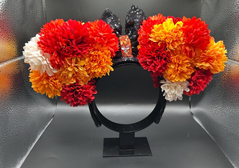 May include: A headband featuring two large circular clusters of artificial flowers in shades of red, orange, and white. Between the floral clusters sits a black sequined bow with a central orange sequined knot featuring a small silver icon.
