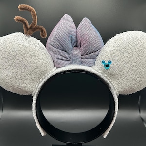 May include: A festive headband featuring white sequined ears, a shimmering bow with blue and purple hues, and a brown pipe cleaner antler. A blue jewel is on one ear. The headband is white with a black interior.